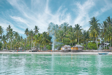 Looking across the water at a fishing village in Papua New Guinea, with a camp fire and people and dogs gathered together on the beach with the huts and fishing boats and palm trees.
